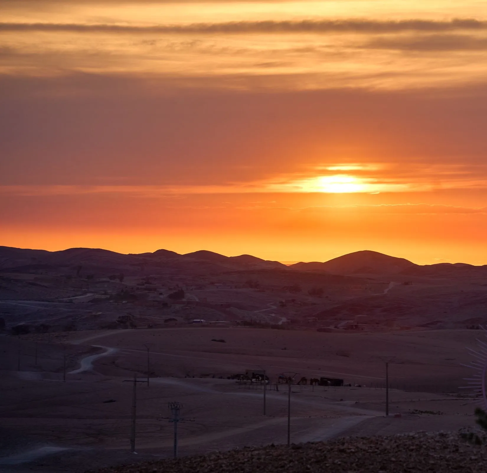 Coucher de soleil sur un désert de pierre marocain : collines sombres en enfilade, ciel orangé et pistes de terre