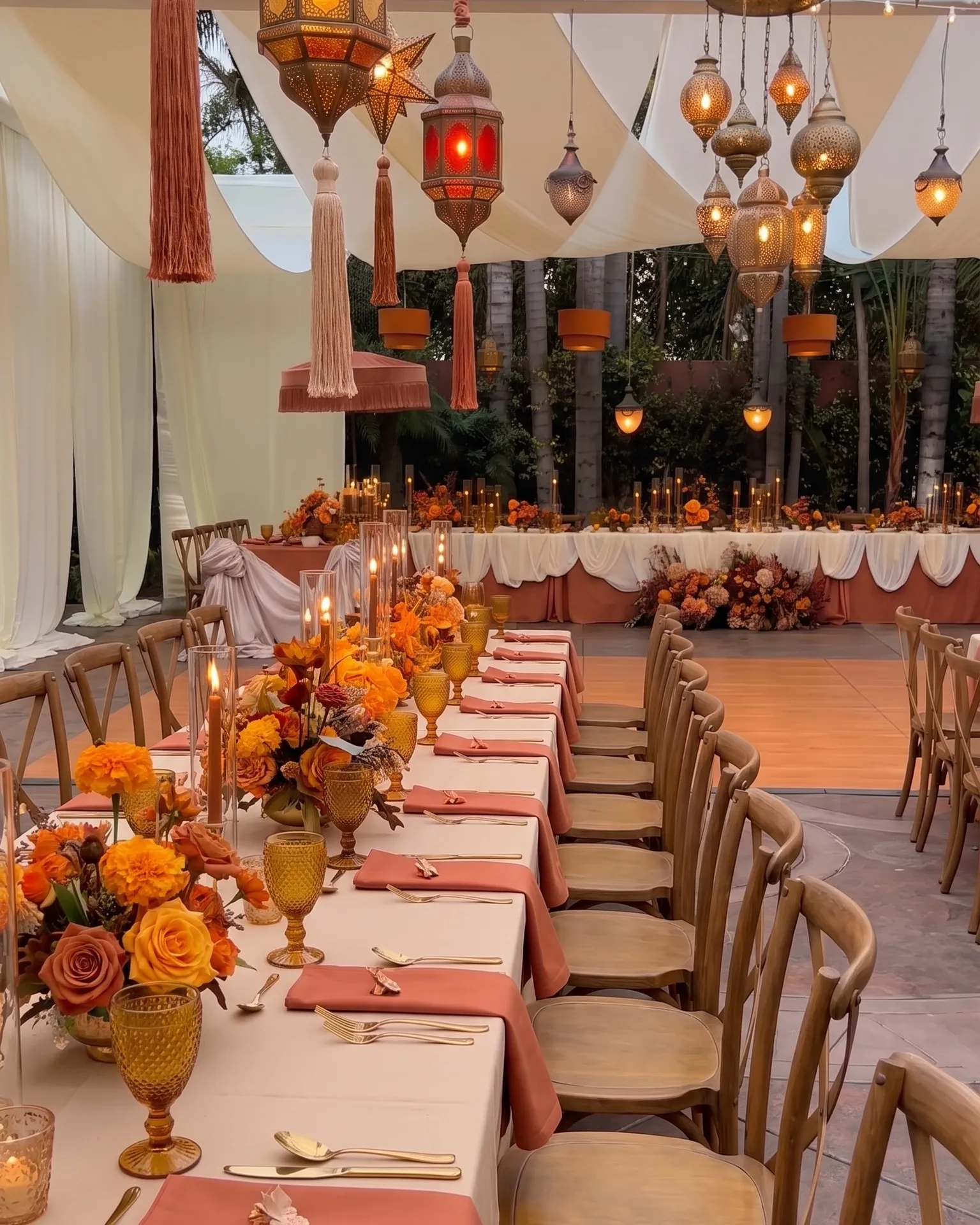 Réception en extérieur sous voiles blancs : lanternes marocaines suspendues et guirlandes lumineuses, longues tables dressées en terracotta et ivoire, bouquets de soucis et de roses orangées