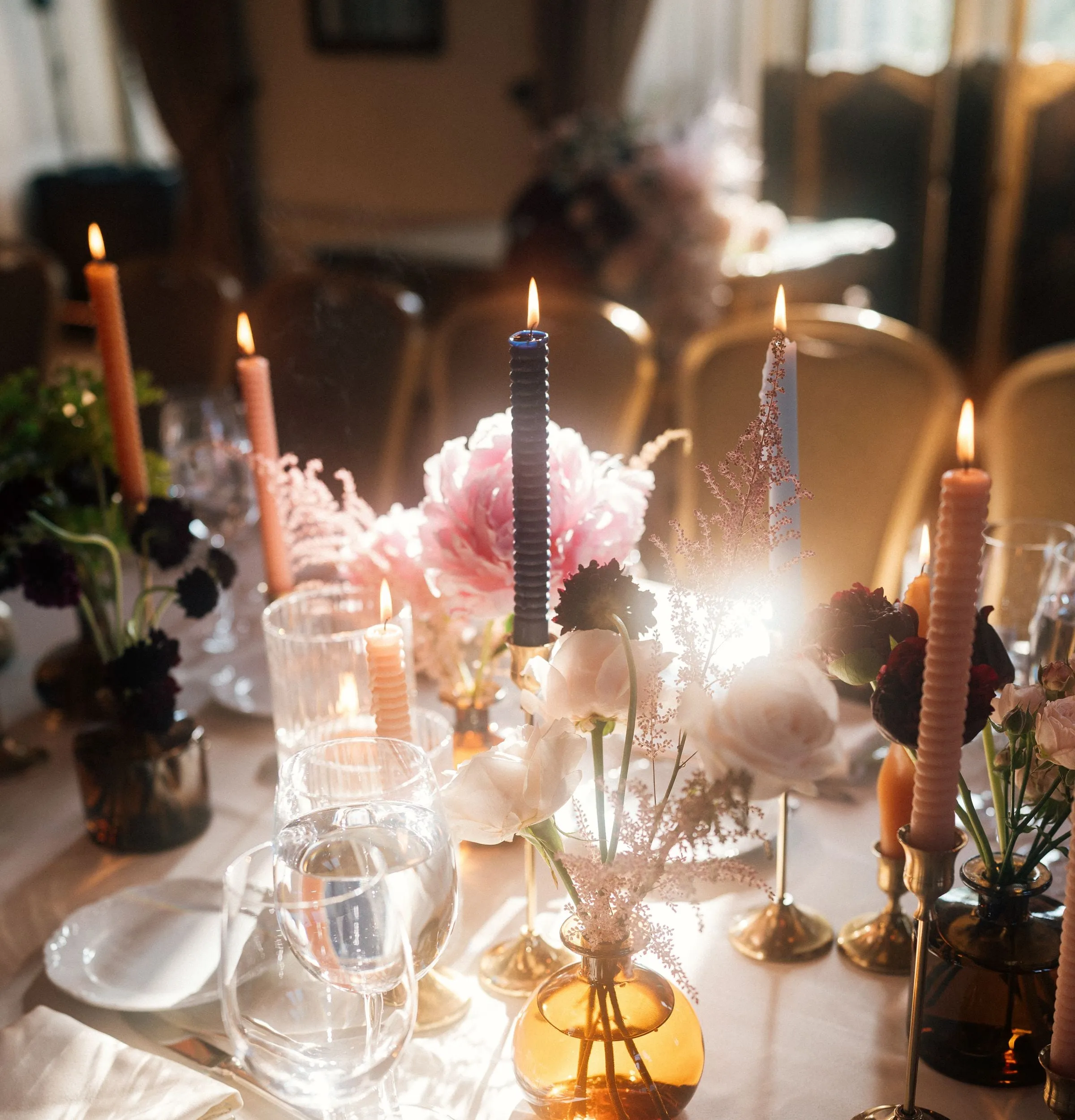 Table de mariage dressée en intérieur à la tombée du jour : bougies torsadées rose et prune, pivoines et roses poudrées, dahlias bordeaux, bougeoirs en laiton et soliflores ambrés