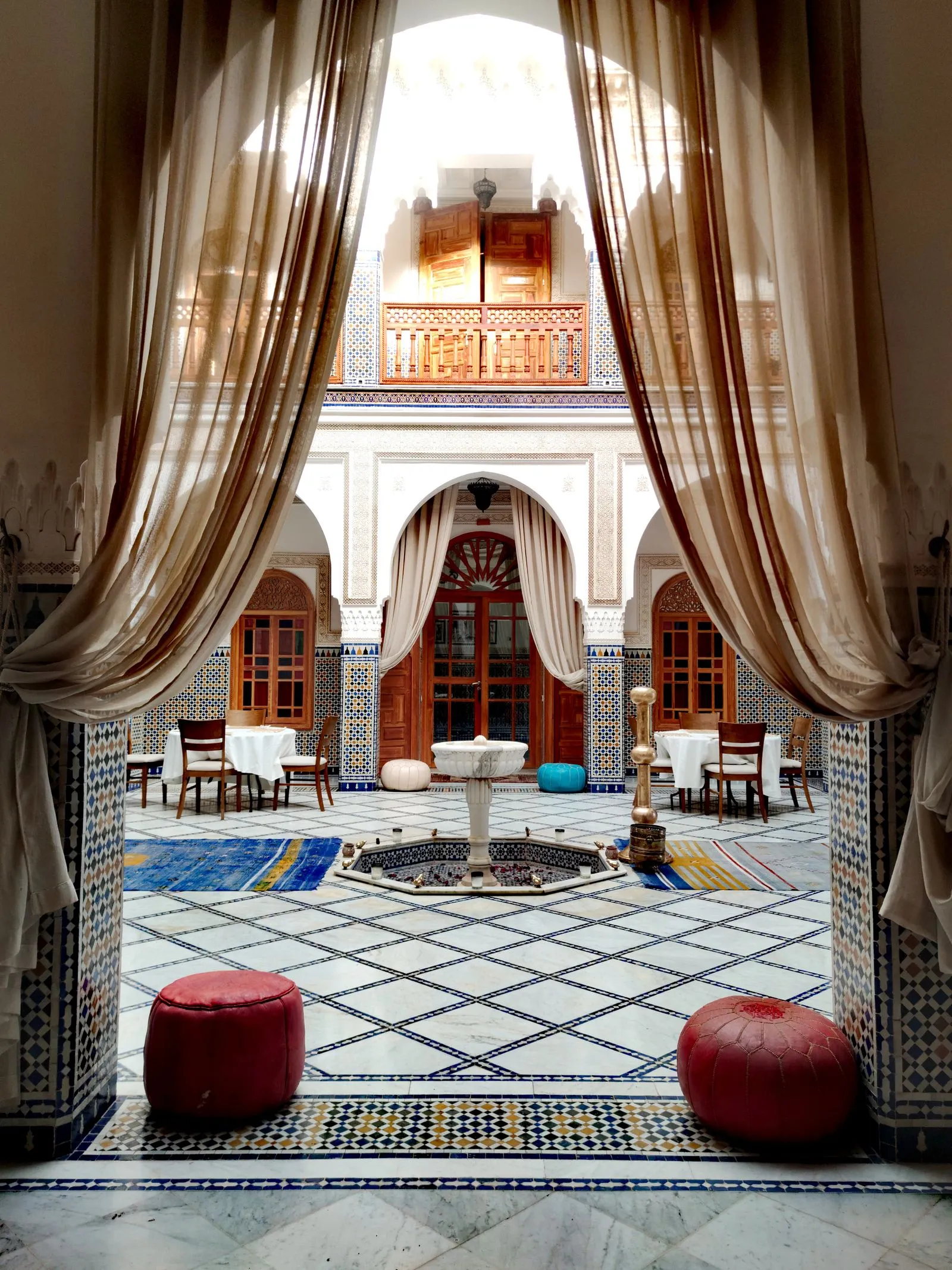 Cour de riad dévoilée par des voilages : arcades, zellige, fontaine et tables dressées