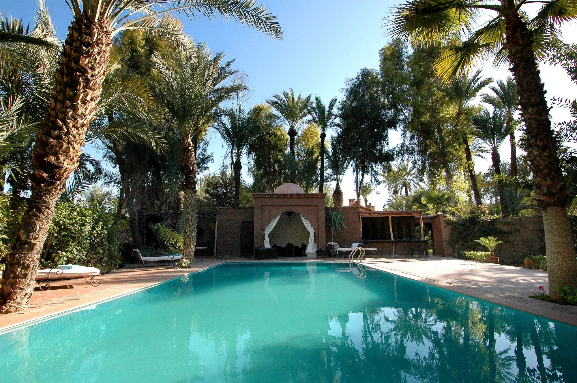 Bassin turquoise bordé de palmiers dattiers, pavillon marocain à coupole et rideaux blancs au fond du jardin