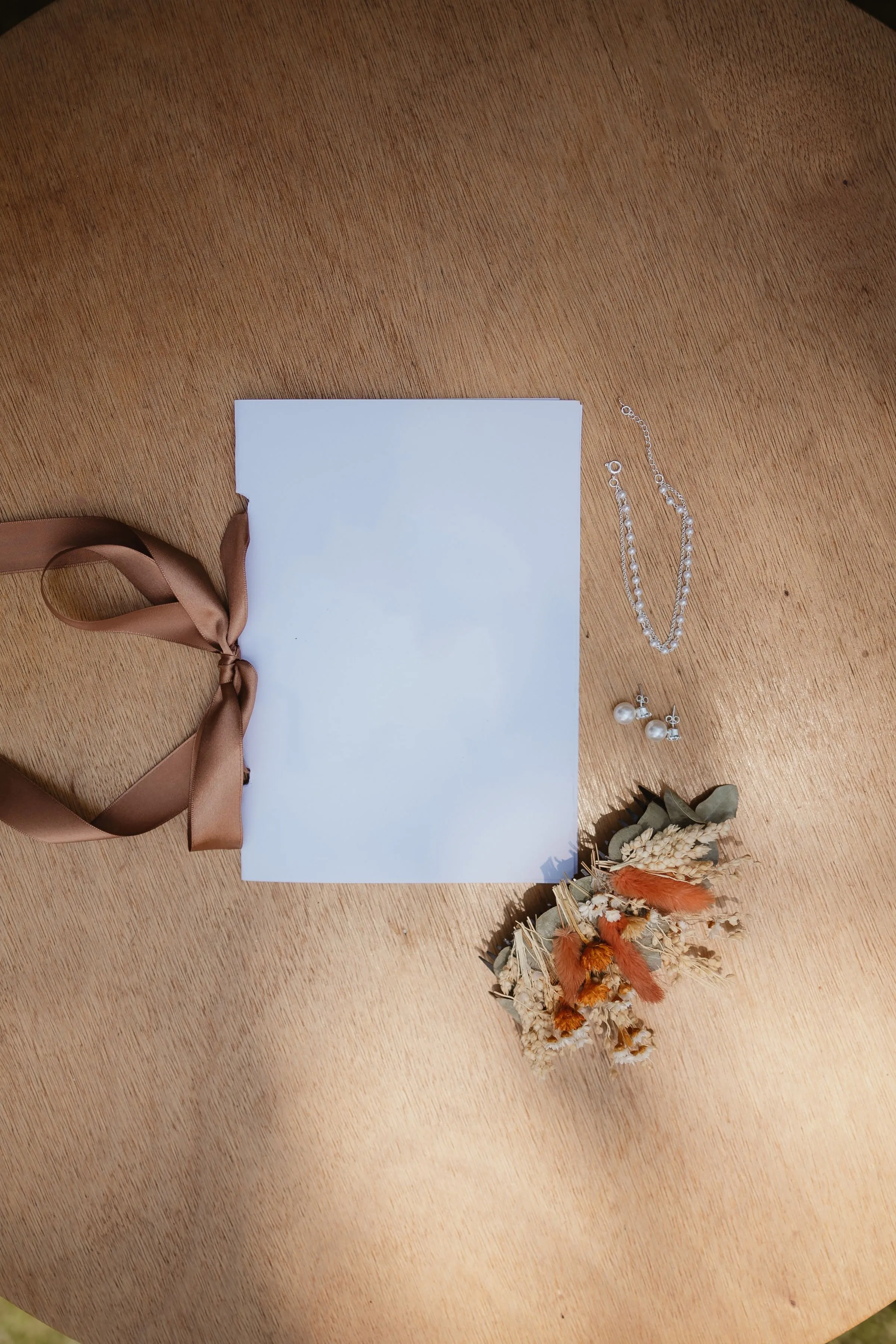 Papeterie de mariage à plat sur une table en bois clair : carte vierge nouée d'un ruban caramel, bouquet de fleurs séchées et bijoux de perles