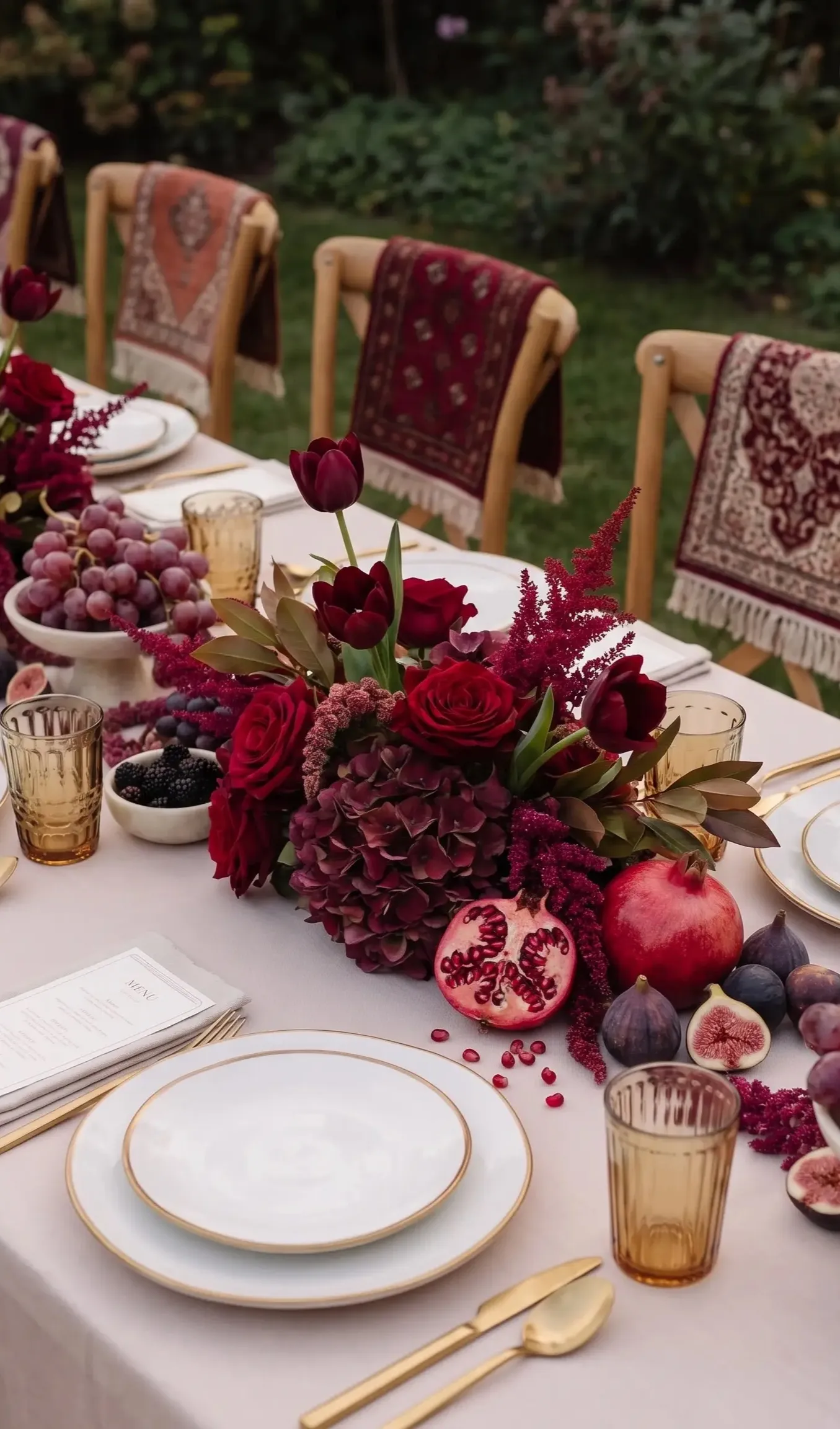 Table de dîner dressée au jardin : hortensias, roses et tulipes bordeaux, grenades ouvertes, figues et raisin, verres ambrés, couverts dorés et tapis berbères posés sur les chaises