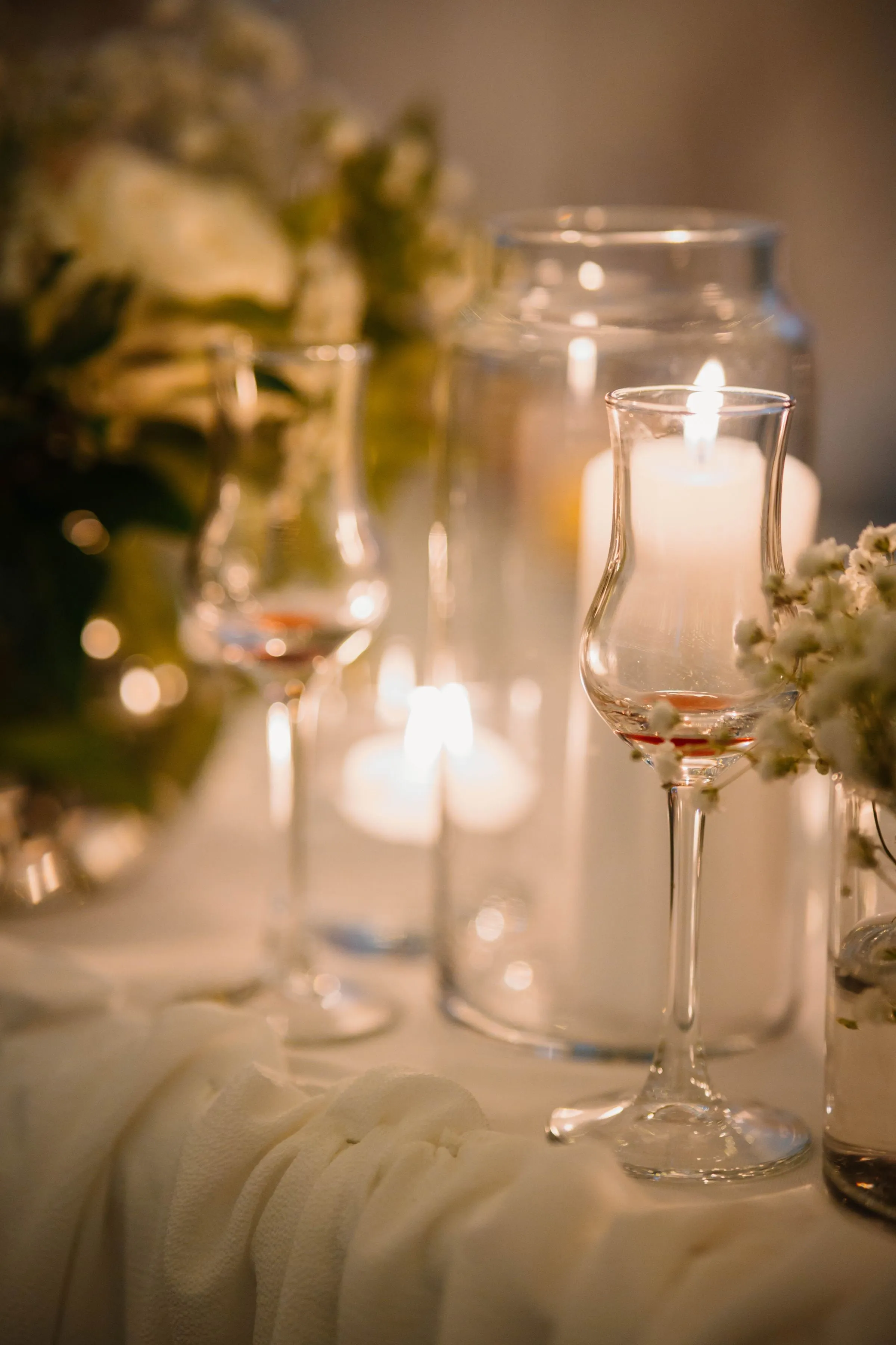 Détail d'une table de réception aux chandelles : verres à pied, bougies flottantes dans leurs photophores de verre, gypsophile et nappe ivoire